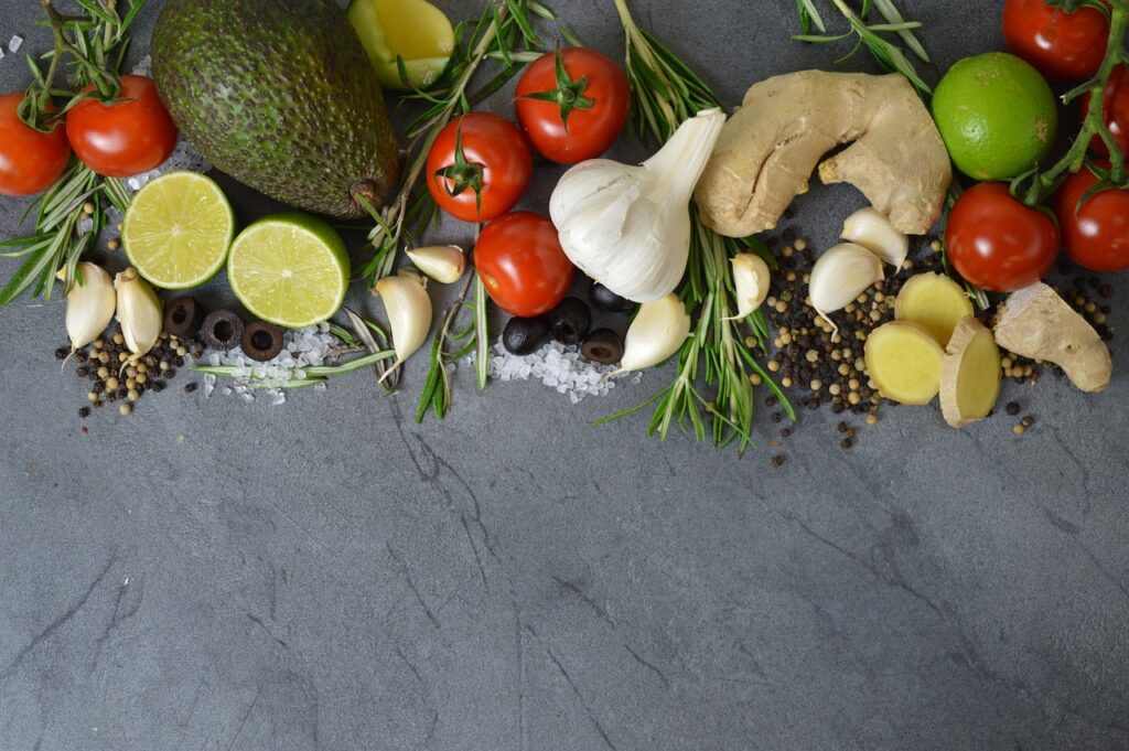 Garlic, avocado, ginger, tomatoes, and greens with spices strewn across a gray background.