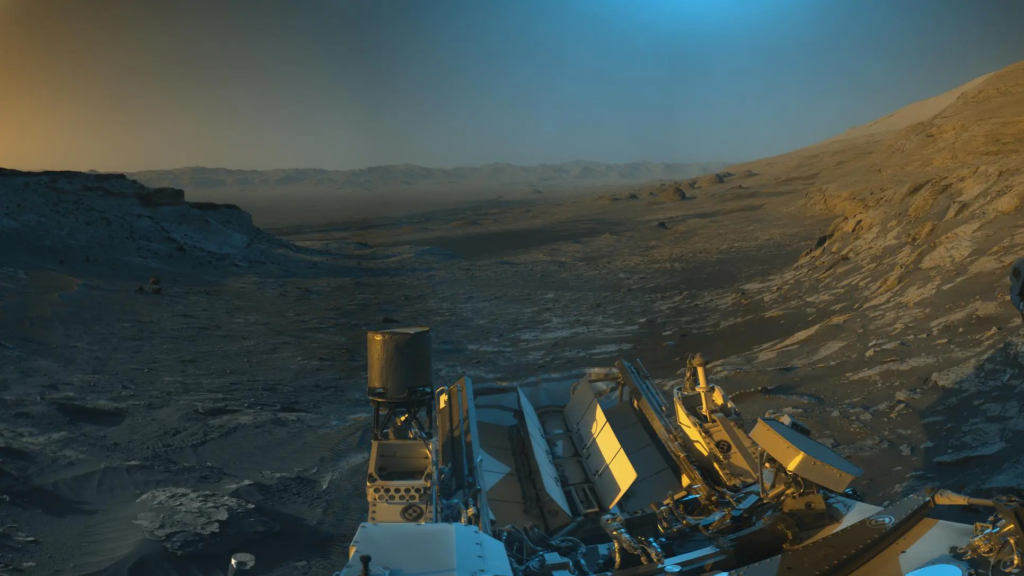 NASA’s Curiosity Mars rover used its black-and-white navigation cameras to capture panoramas of this scene at two times of day. Blue, orange, and green colors were added to a combination of both panoramas for an artistic interpretation of the scene.
