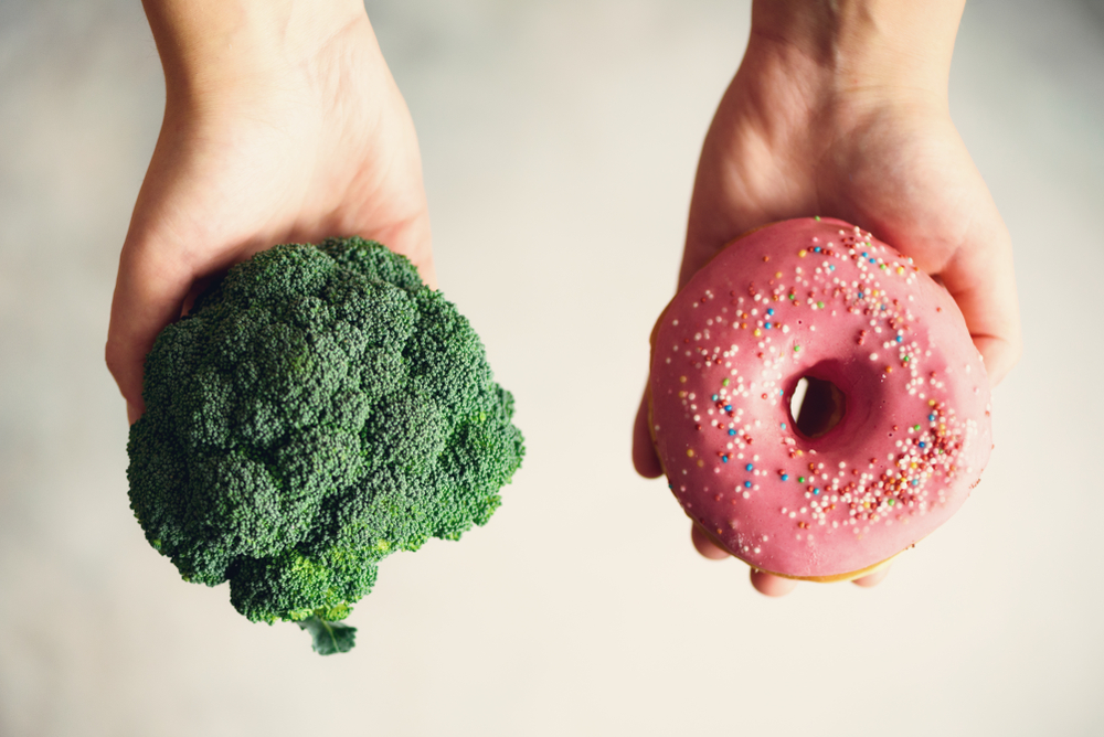 Young woman in white T-shirt choosing between broccoli or junk food, donut. Healthy clean detox eating concept. Vegetarian, vegan, raw concept. Copy space.