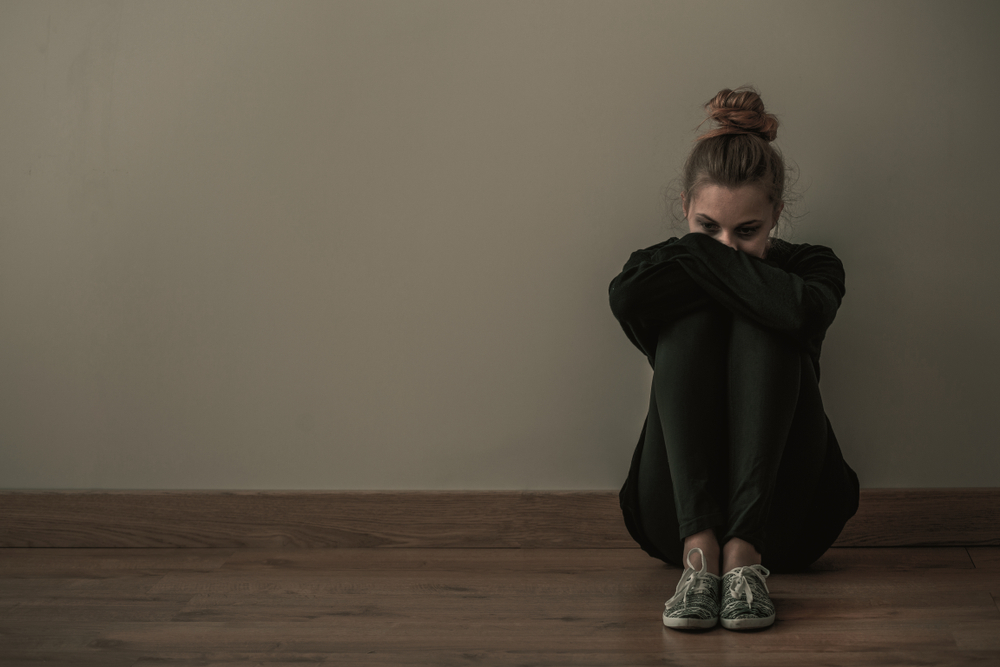Young woman with anxiety disorder wearing dark clothes sitting on the floor, copy space on empty wall