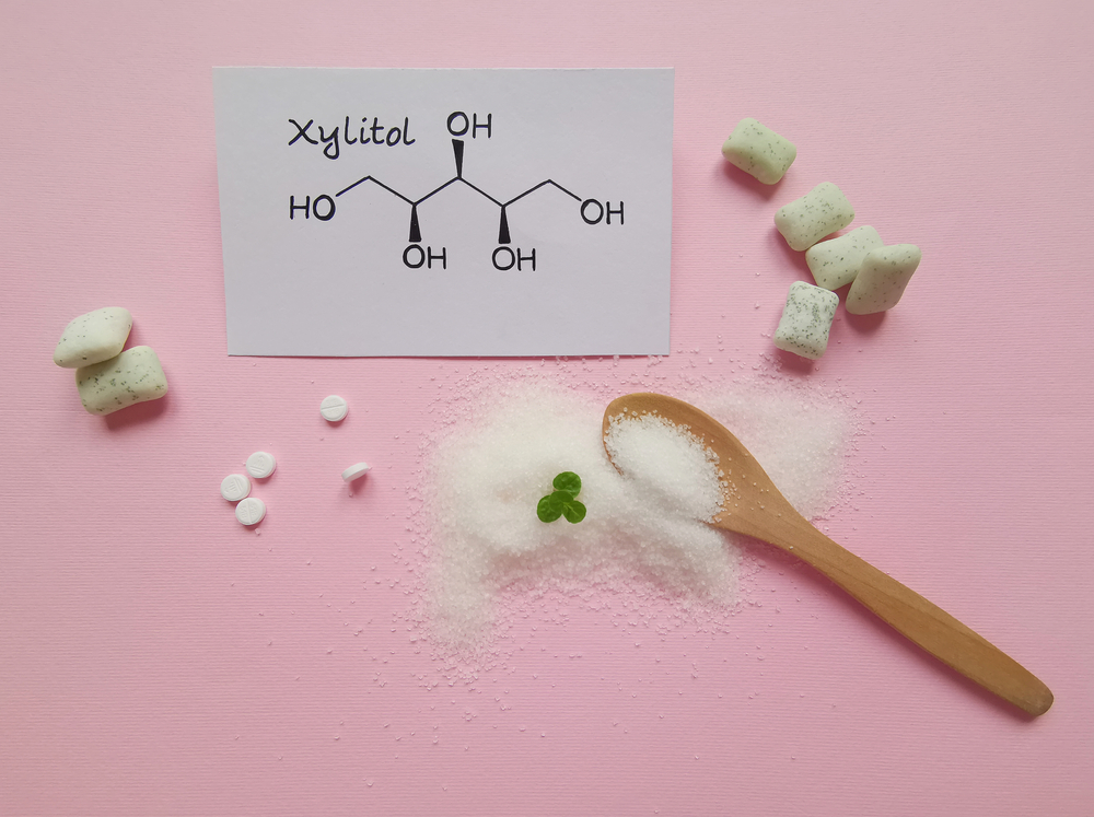Structural chemical formula of xylitol molecule with chewing gum in the background. Xylitol is a polyalcohol (sugar alcohol), used as a food additive and sugar substitute in toothpaste and chewing gum