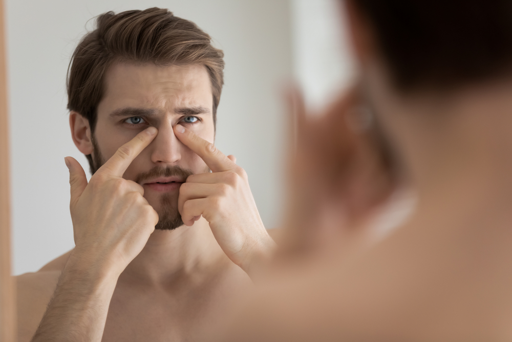 Head shot close up unhappy young man touching under eyes area or facial skin, feeling dissatisfied with condition, looking in mirror after morning showering, needs improving skincare daily routine.