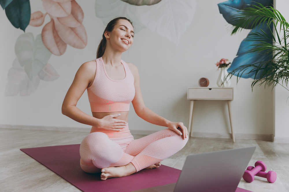 Full body fun satisfied happy smiling young sporty fitness trainer instructor woman in pink tracksuit do yoga exercise use laptop pc computer close eyes hold tummy sit on mat floor at home gym indoor