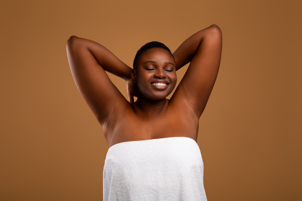 Portrait Of Happy Chubby Black Female Posing With Hands Up, Showing Her Smooth Armpits, Cheerful African American Lady With Beautiful Skin Posing In White Towel Over Brown Studio Wall, Copy Space
