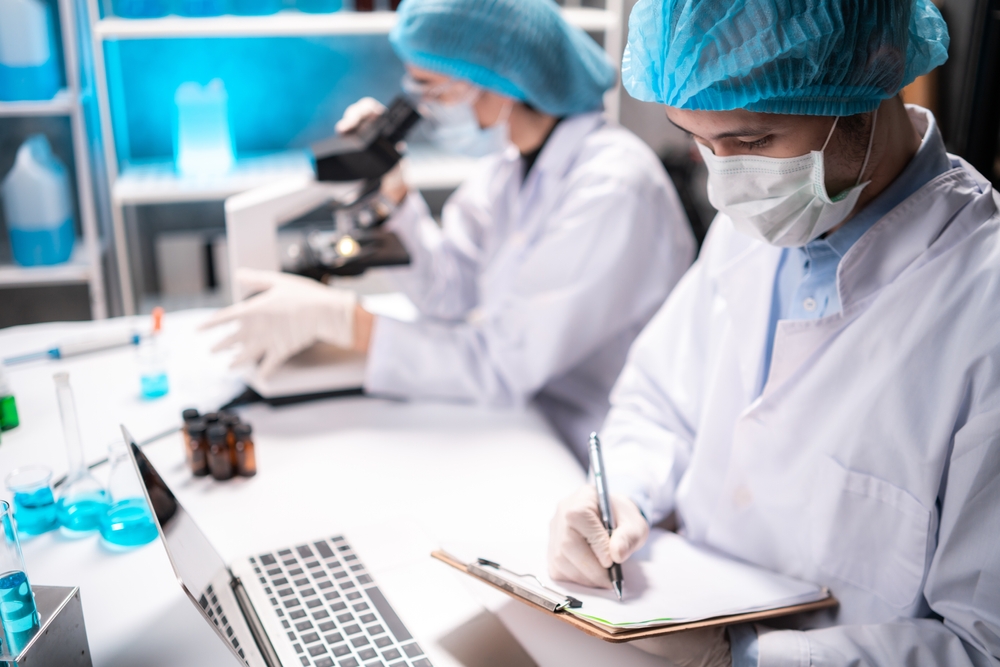Medical healthcare researcher and scientist writing experiment detail on paper in clipboard while looking at laptop for details with colleague looking through microscope in background in laboratory