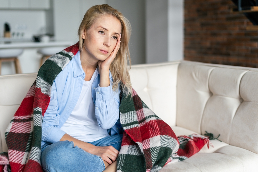Ill and exhausted woman touch forehead, having headache, chronic migraine or depression, sitting on couch with blanket. Upset frustrated female feel stressed, fever or high blood pressure in temples