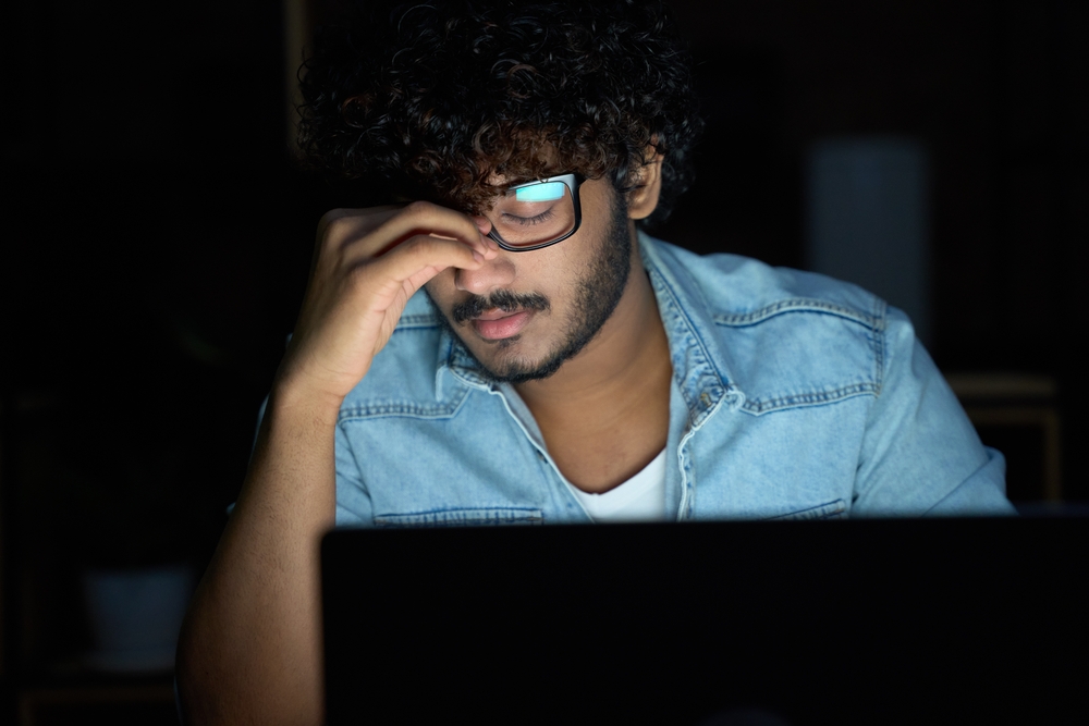 Tired sleepy stressed overworked young indian business man student wearing glasses feeling lack of sleep, having eyestrain vision problem, using laptop computer working late at night at home office.
