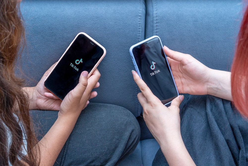 Two teenage girls with phones in their hands are sitting at home on bed. Social Media tiktok is loaded on screen.
