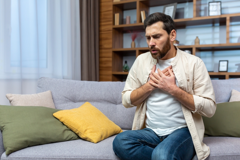 A young man is sitting on the sofa at home and holding his chest. Feels severe pain, shortness of breath, panic attack, heart attack, stroke.
