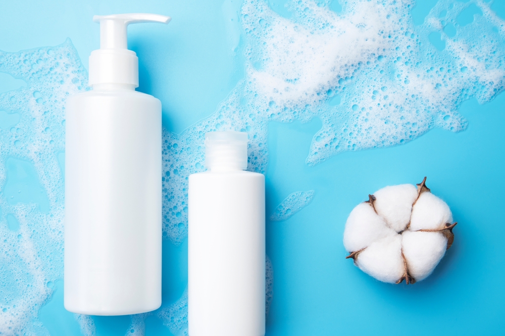 Natural organic cosmetics with cotton extract. White cosmetic tubes, soap foam, cotton flowers on blue background. Face wash mousse soap shampoo shower gel. Beauty spa skin care mockup