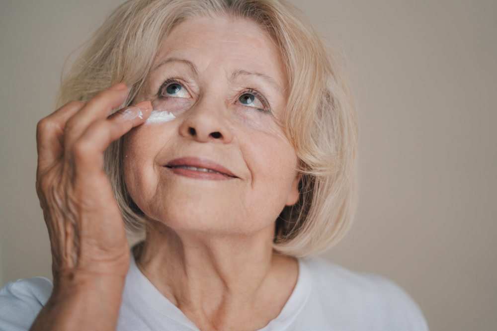 Smiling old caucasian woman applying anti aging cream on her face against grey background. Beauty portrait. Facial treatment. Anti-wrinkles care. Anti-aging
