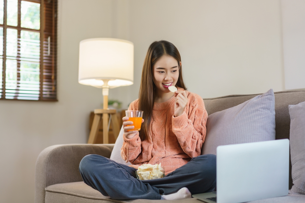 Women drinking orange juice and eating snack while watching movie on laptop in lifestyle at home.