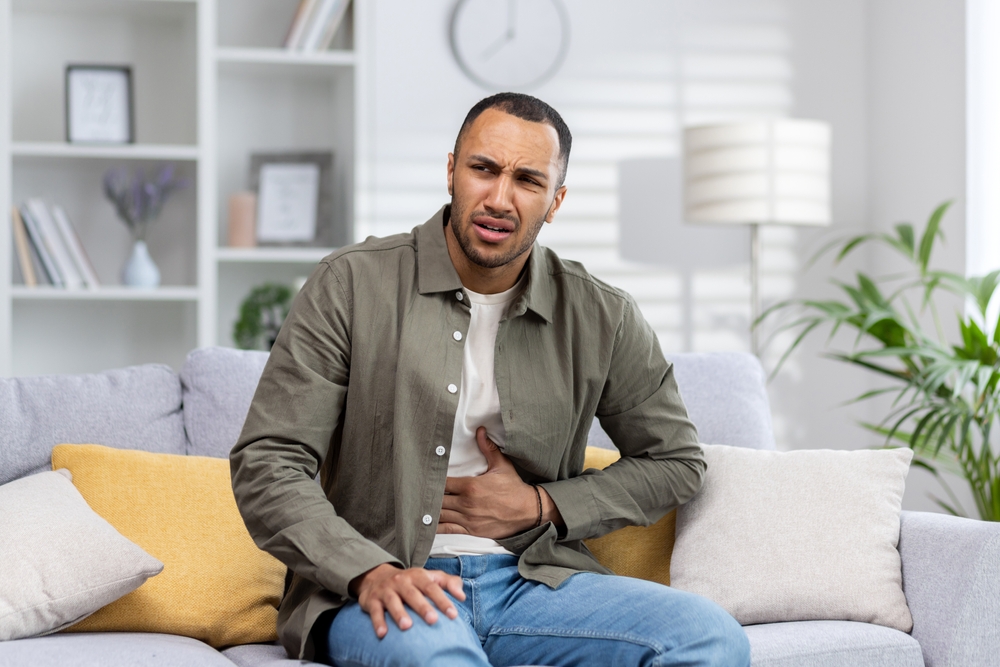 A young African American man is suffering from abdominal pain, stomach cramps and constipation. He is sitting on the sofa and holding his stomach, grimacing in pain.
