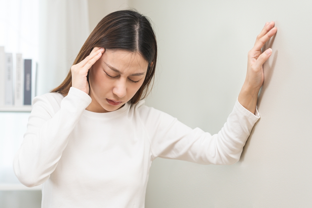 Dizzy asian young woman, girl headache or migraine pain. suffering from vertigo, difficulty standing up leaning on wall at home, holding head with hand, health problem of brain, inner ear not balance.

