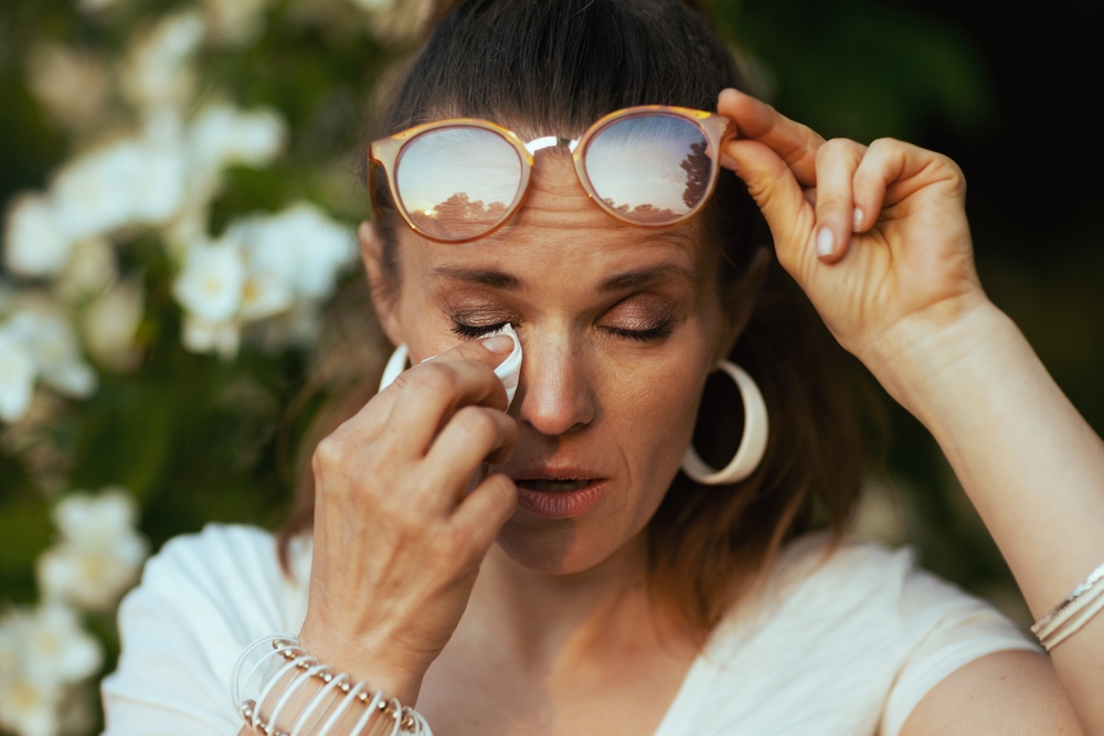 Summer time. sad young female in white shirt with handkerchief and eyeglasses has an allergy attack near flowering tree.