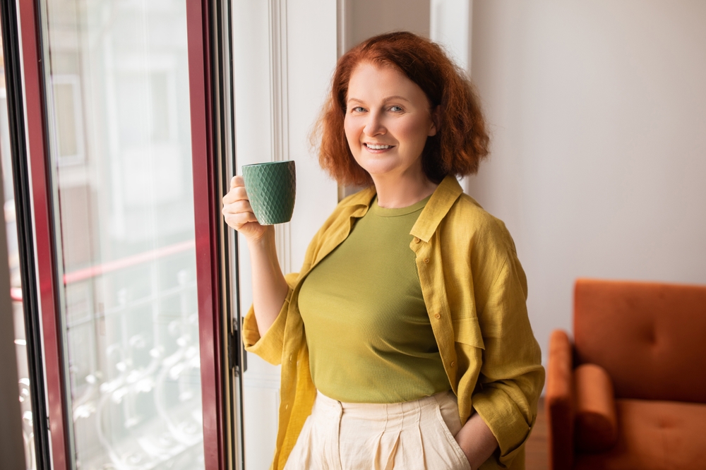 Smiling Senior Woman With Cup Of Tea Standing Near Window At Home, Happy Older Lady Enjoying Hot Drink And Looking At Camera, Beautiful Retired Female Relaxing In Cozy Interior, Free Space