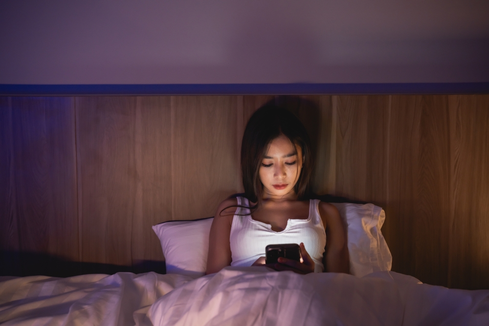 Asian woman using mobile phone smartphone laying on the bed in the bedroom. Sleepy exhausted, can not sleep. Insomnia, addiction concept. Women scrolling social networks on mobile dark bedroom.