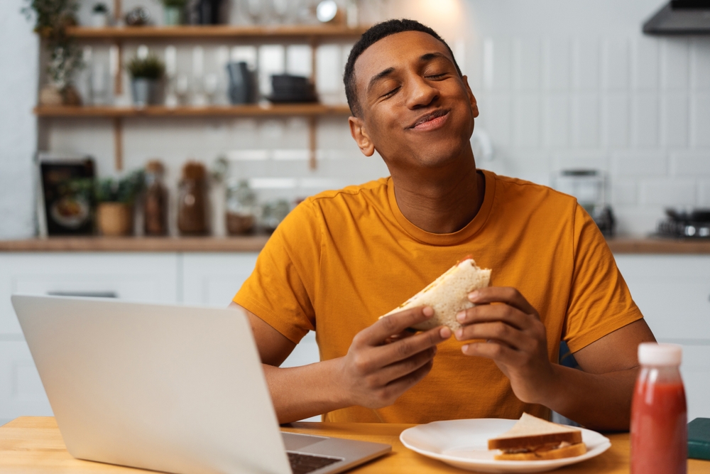 Handsome, happy Latin man holding tasty sandwich, eating, enjoying with closed eyes sitting in modern kitchen at home. Concept of food, freelancer working at home