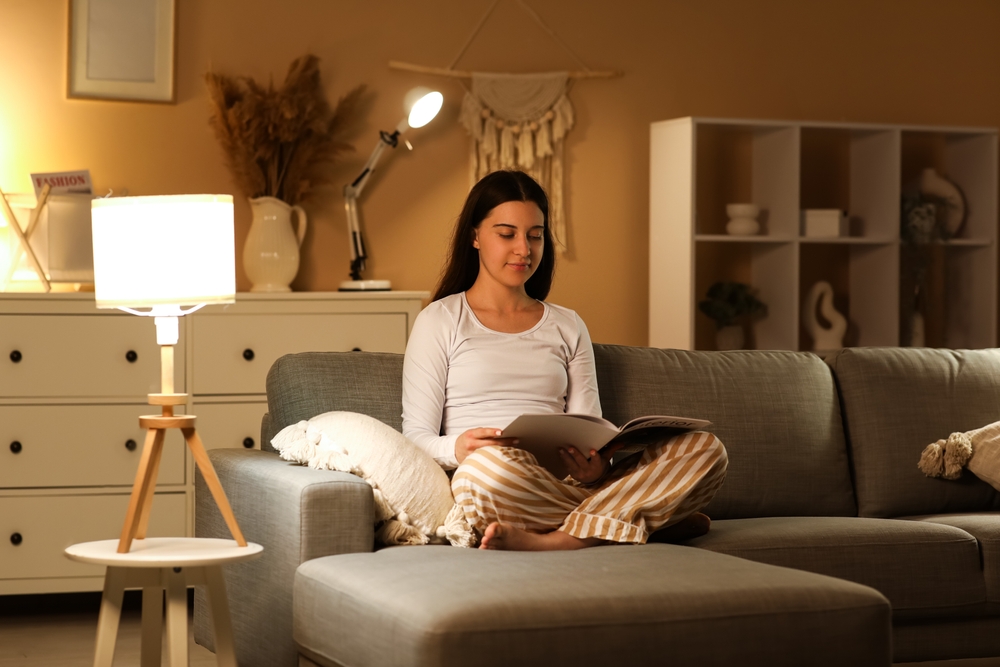 Young woman in pajamas reading magazine on soft couch in living room at night