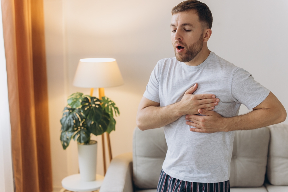 A young man is standing at home and holding his chest. Feels severe pain, shortness of breath, panic attack, heart attack, stroke.
