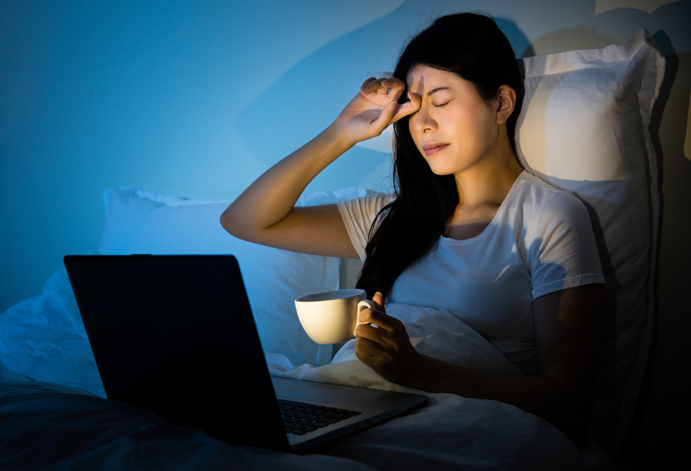 businesswoman rubbing eyes drinking coffee to working with computer sitting on bed at room. mixed race asian chinese model.