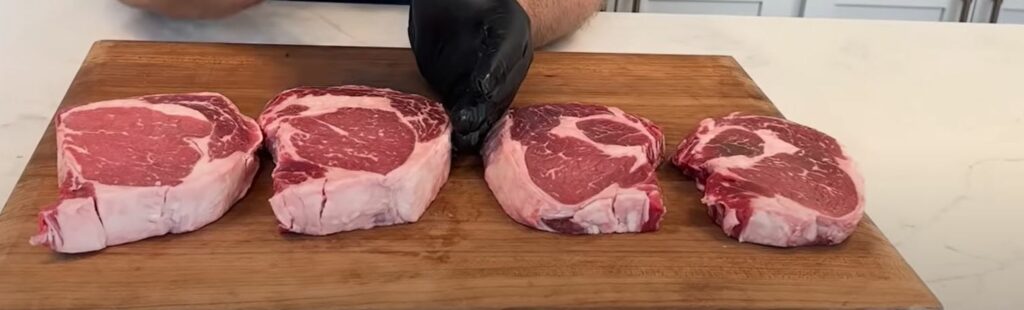 Four steaks on a wooden cutting board.