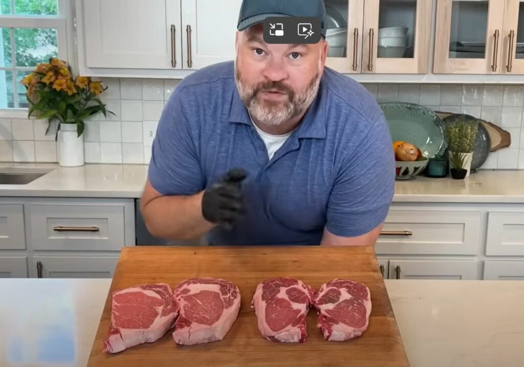 A man speaking about steaks on a cutting board. Kitchen cabinets and countertops in the background.
