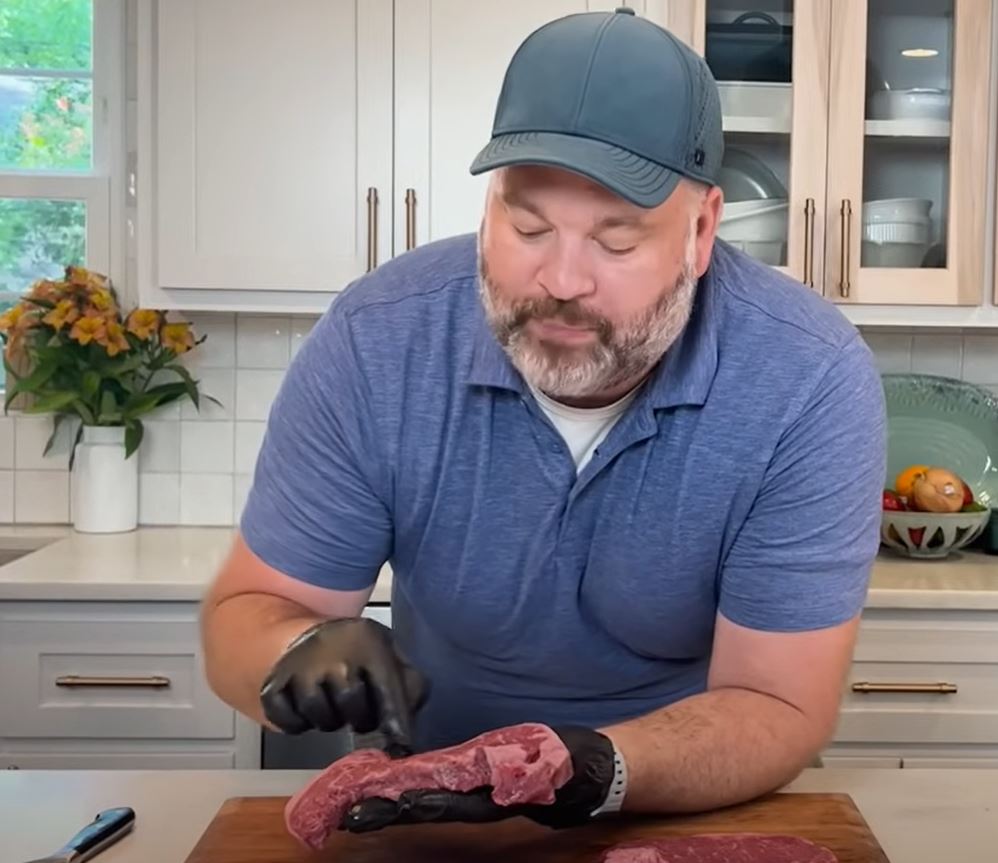 A man seasoning a cut of steak. Kitchen counter and cabinets in the background.
