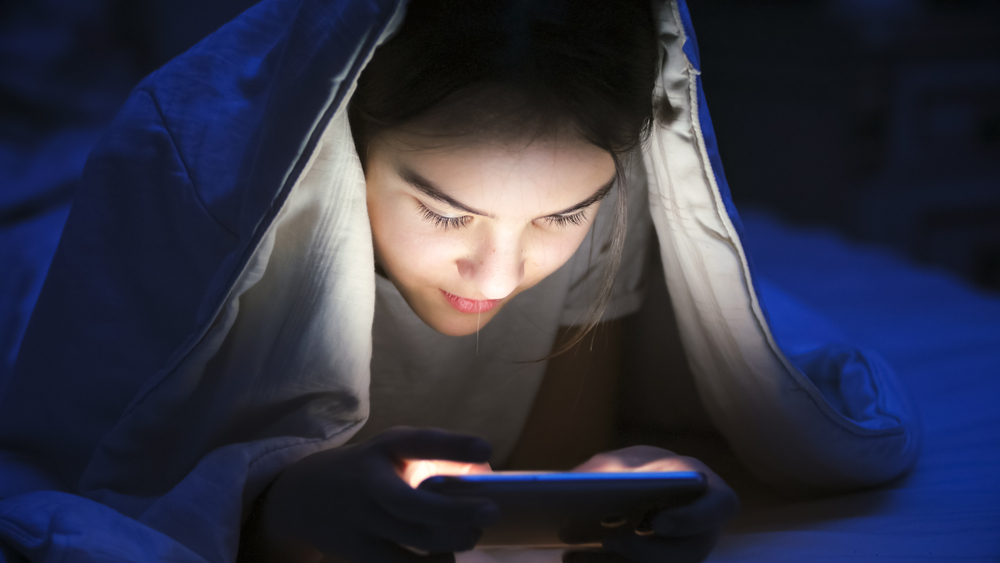 Portrait of smiling girl with mobile phone under blanket at night
