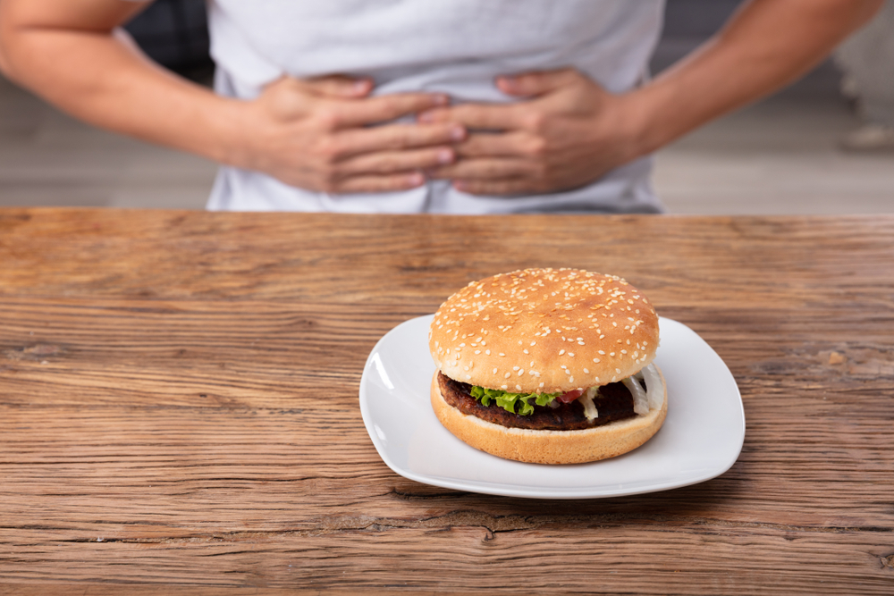 Man Having Stomach Pain Near Fresh Burger On Plate Over Wooden Desk