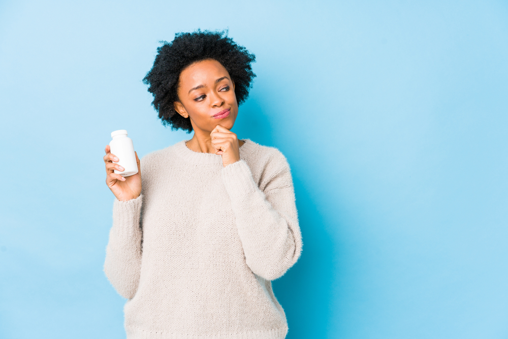 Middle age african american woman holding a vitamin bottle looking sideways with doubtful and skeptical expression.

