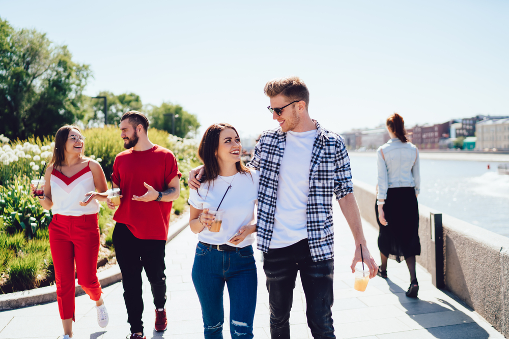 Caucasian male and female friends walking at city embankment enjoying live meeting for communicate, happy hipster guys with non alcohol cocktails to go spending leisure weekend togetherness