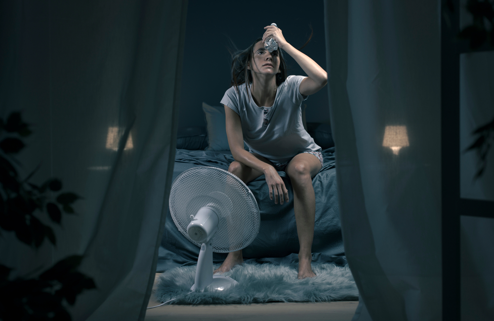 Exhausted woman suffering suring the heatwave, she is holding a water bottle and sitting in front of a cooling fan in the bedroom