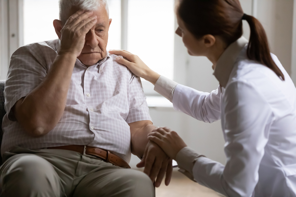 Unhealthy mature old patient complaining to doctor about head ache migraine illness at checkup meeting in clinic. Professional young female general practitioner giving advice, treating elderly man.
