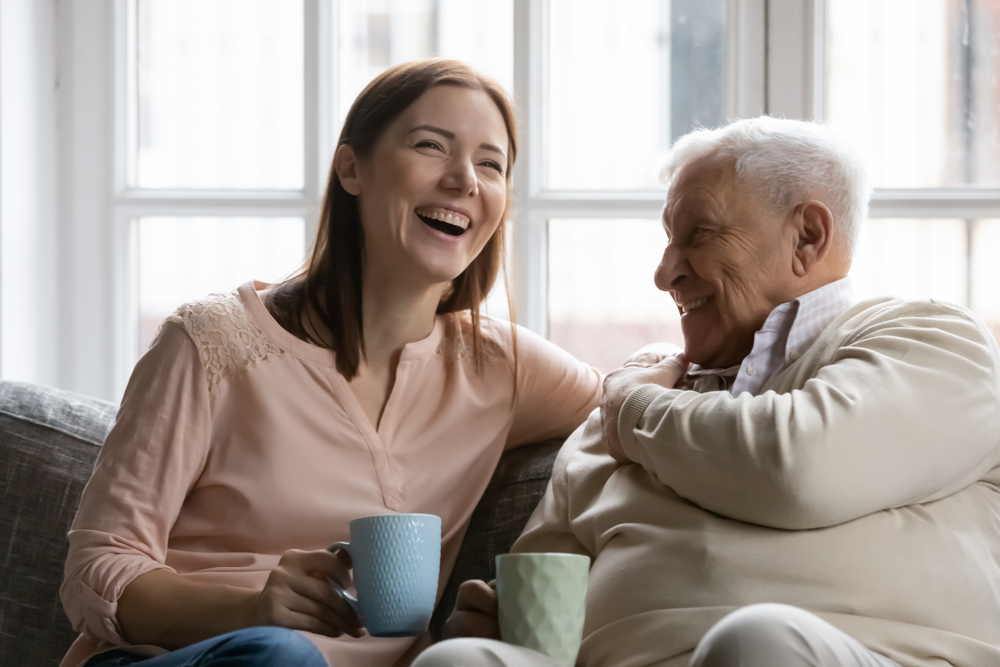 Family members of two generations grown daughter and mature father having fun enjoy tea talk on cozy couch. Attentive young lady caregiver social worker visit support care for glad elderly man patient

