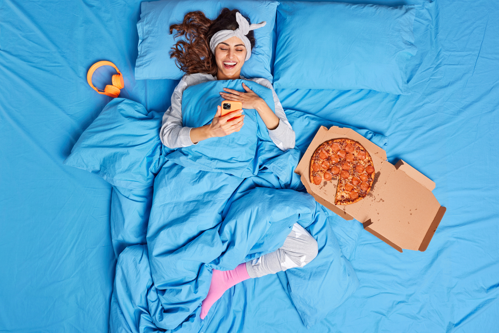 Positive brunette European woman enjoys video call in bed holds modern smartphone lies under soft blanket eats pizza at bedroom has lazy day enjoys cozy domestic atmosphere. Bedtime concept.