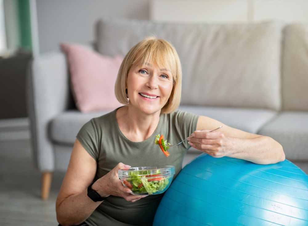 Exercise and healthy diet concept. Senior woman with fitball eating fresh vegetable salad at home. Mature Caucasian lady having veggies after sports training, keeping weight loss diet
