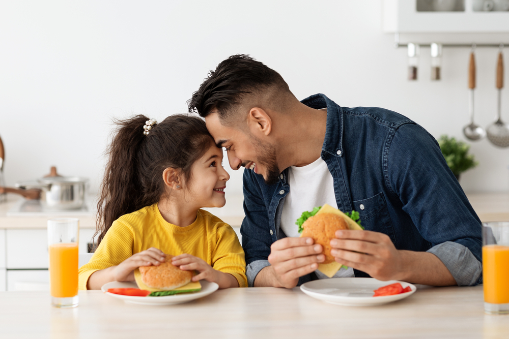 Happy Loving Arab Dad And Daughter Eating Sandwiches In Kitchen And Bonding, Cheerful Middle Eastern Father And Cute Little Female Child Touching Foreheads And Smiling, Enjoying Time Together
