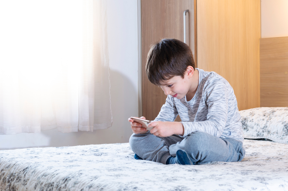 Little 6 years old boy using and playing on smarphone. Cute little boy playing games on smartphone in bed. Boy plays in video game on his smartphone. Social and technology concept.
