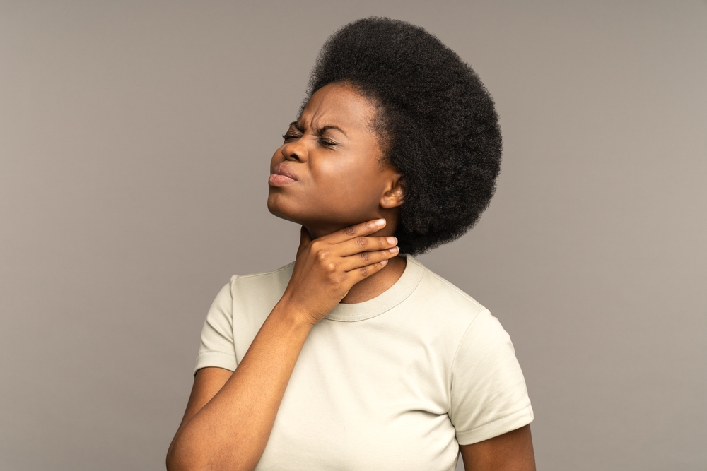 Unhealthy african girl having sore throat, suffering from painful swallowing, strong pain in throat or loss of voice holding neck isolated on studio grey background. Tonsillitis angina thyroid concept