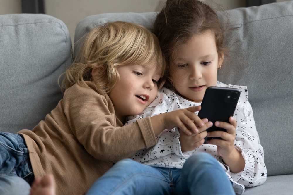 Two little toddler preschooler sibling gen Z kids using online parental control app on mobile phone, playing video game for children, sharing digital gadget, relaxing on comfortable couch at home
