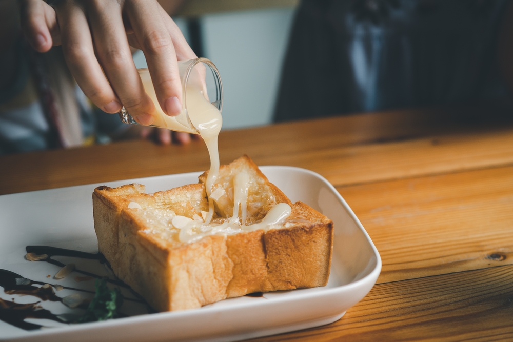 Woman pouring condensed milk on toast on the dining table, woman eating toast in the cafe restaurant. Food bakery concept.
