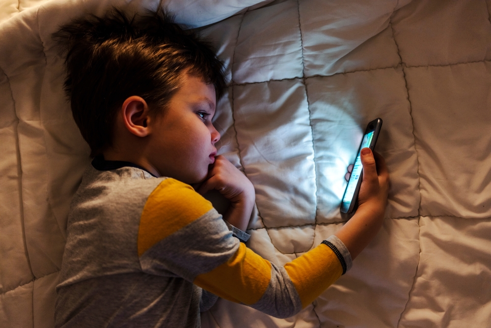 Cute little boy using a smartphone. Kid playing with mobile phone, lying on a bed. Freetime. Technology and internet concept. Smiling toddler laying on bed and looking at smartphone. Light reflectio
