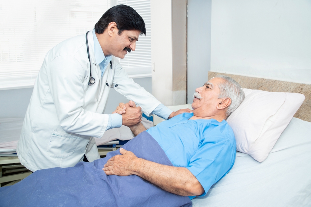 Smiling Indian caring doctor supporting putting hand on shoulder of olde senior male patient lying on bed at clinic or hospital. Elderly people health care concept.