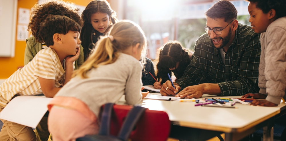 Group of elementary school kids stand around a table in a classroom, attentively following a lesson being taught by their teacher. Young students engaging in a fun activity of colouring and drawing.
