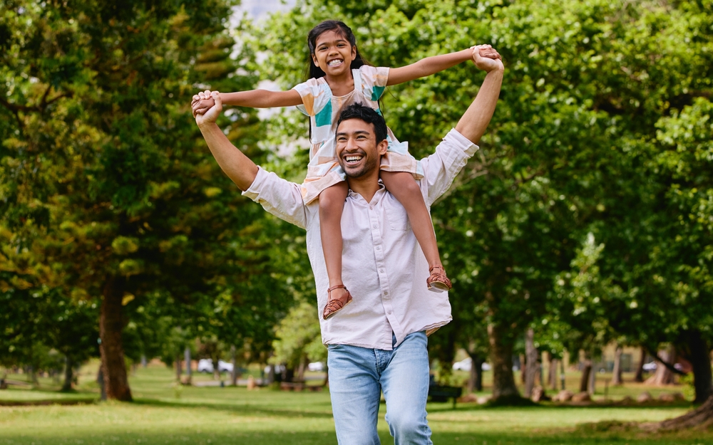 Indian dad, daughter and shoulders in park with smile, airplane game or piggyback in nature on holiday. Man, girl and playing together in garden, woods and summer sunshine for happy family vacation
