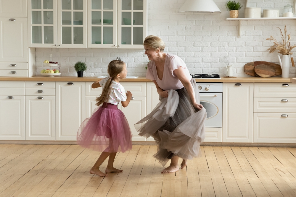 Full length happy barefoot 7s cute little kid girl and older retired grandmother wearing puffy skirts, dancing together in modern kitchen, active different generations family playing on weekend.
