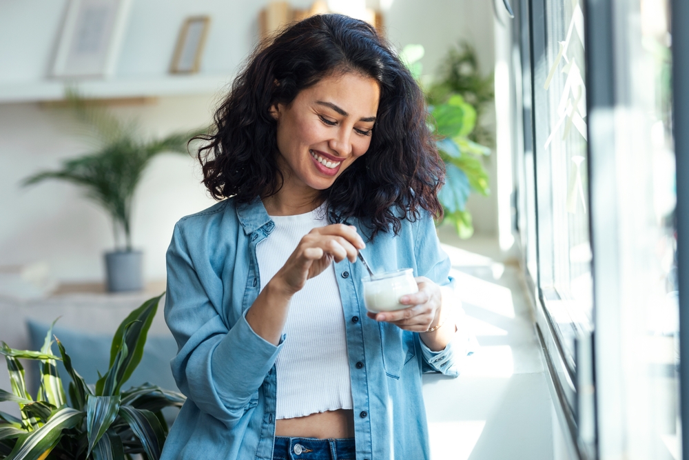 Shot of happy beautiful woman eating yogurt while standing in living room at home.
