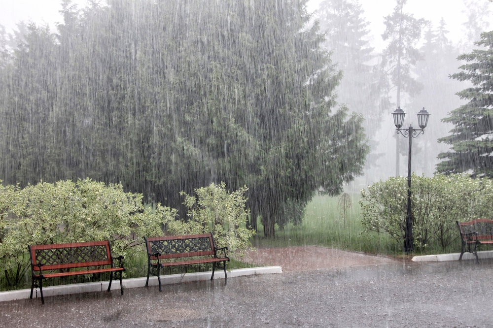 Rain with hail on a summer day. Large puddles on the sidewalk during heavy rain. Heavy rain in the park. Wet benches on a paved path. Spray drops and bubbles in large puddles.
