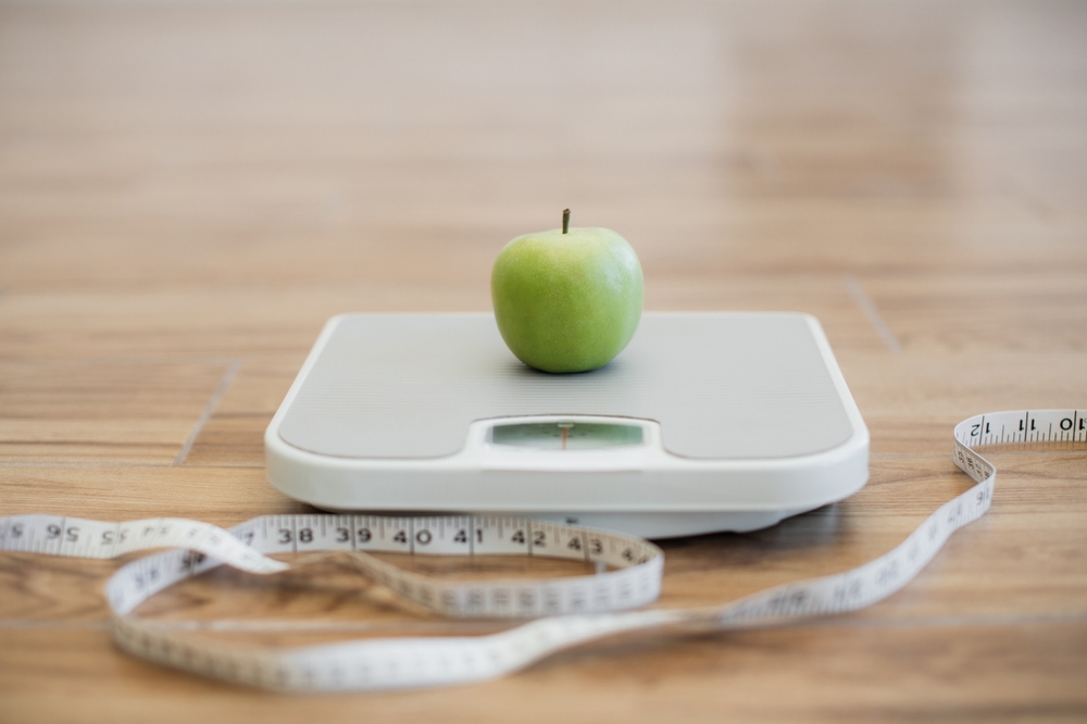 Close up view of green ripe apple being placed on bathroom scales while measuring tape lying on wooden floor nearby. Keeping active and eating balanced diet helping in maintaining healthy weight.